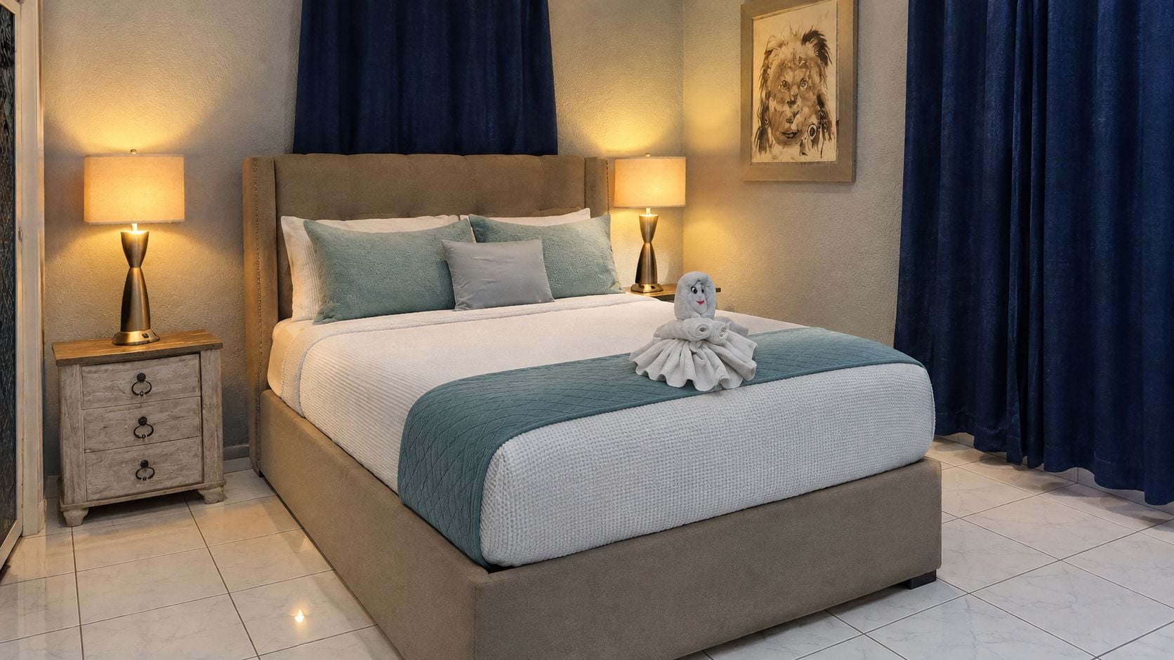 Una cama bien hecha con una colcha blanca y aguamarina, dos almohadas y una toalla con forma de pájaro sobre la cama. Hay dos mesitas de noche con lámparas, una a cada lado de la cama.