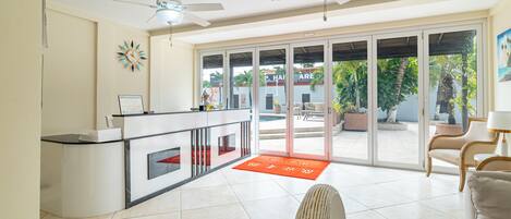 A spacious reception area with a modern design, featuring a reception desk, two ceiling fans, and a large glass door leading to an outdoor area with plants and a view of a building in the background.