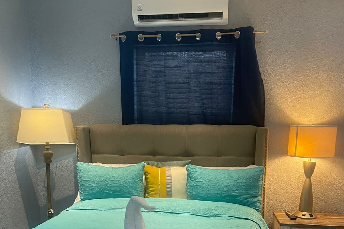 A hotel room with a bed, two bedside lamps, a nightstand, a wall-mounted air conditioner, and a blue curtain.