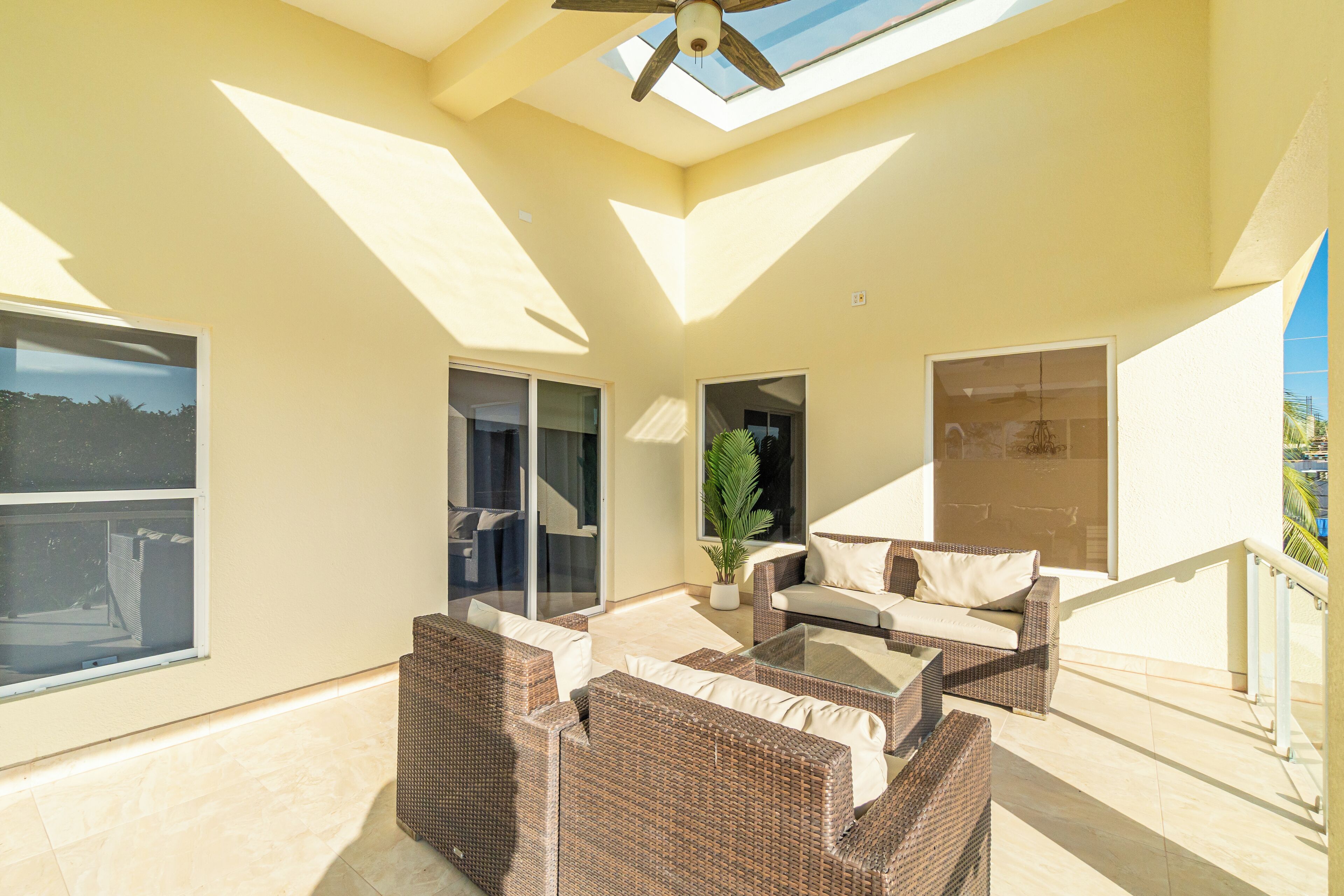 A sunlit patio with wicker furniture, a glass table, and a ceiling fan.