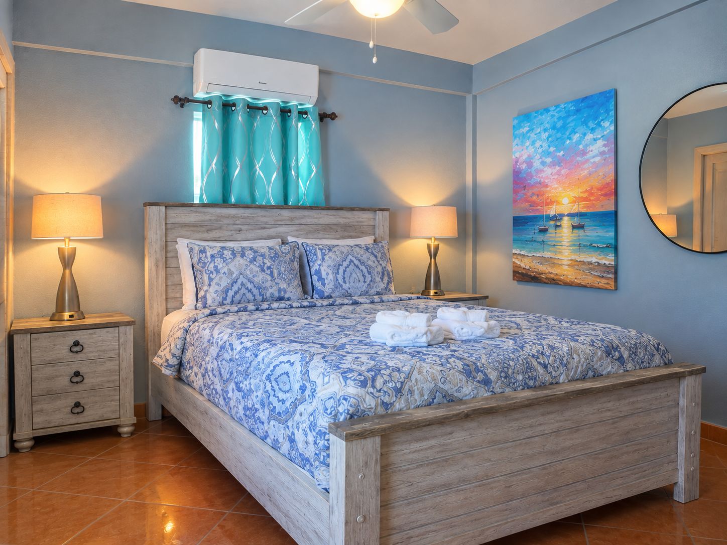 A bedroom with a wooden bed, patterned bedding, a ceiling fan, a painting of a sunset, and a bathroom mirror.