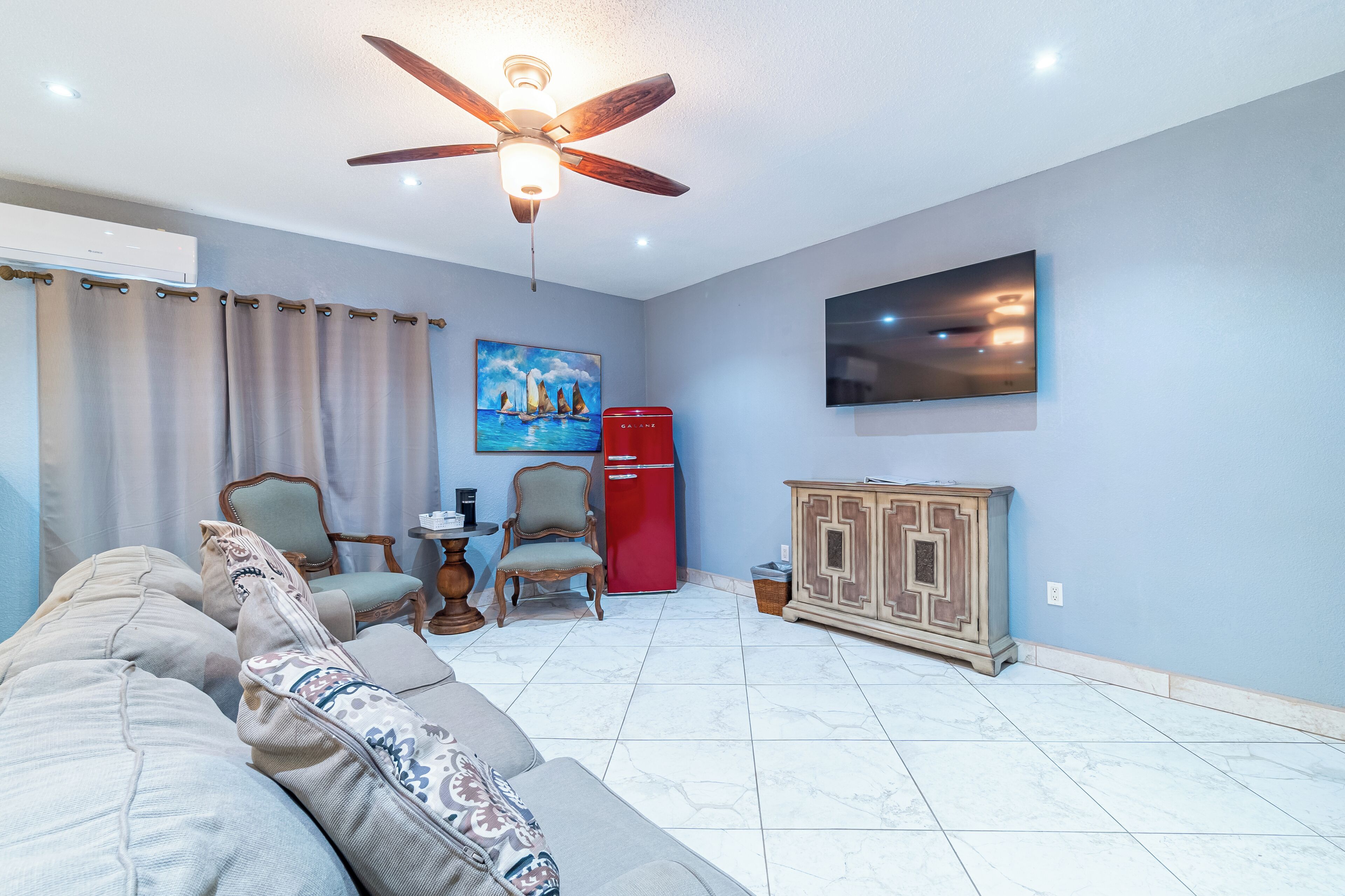 A living room with a ceiling fan, a red refrigerator, a TV mounted on the wall, and a beige sofa with patterned cushions.