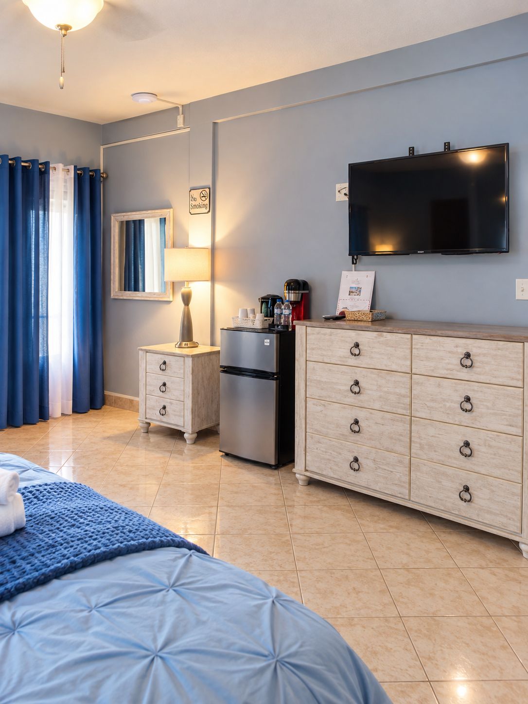 A hotel room with a bed, a TV, a minibar, a coffee machine, and a 'No Smoking' sign.