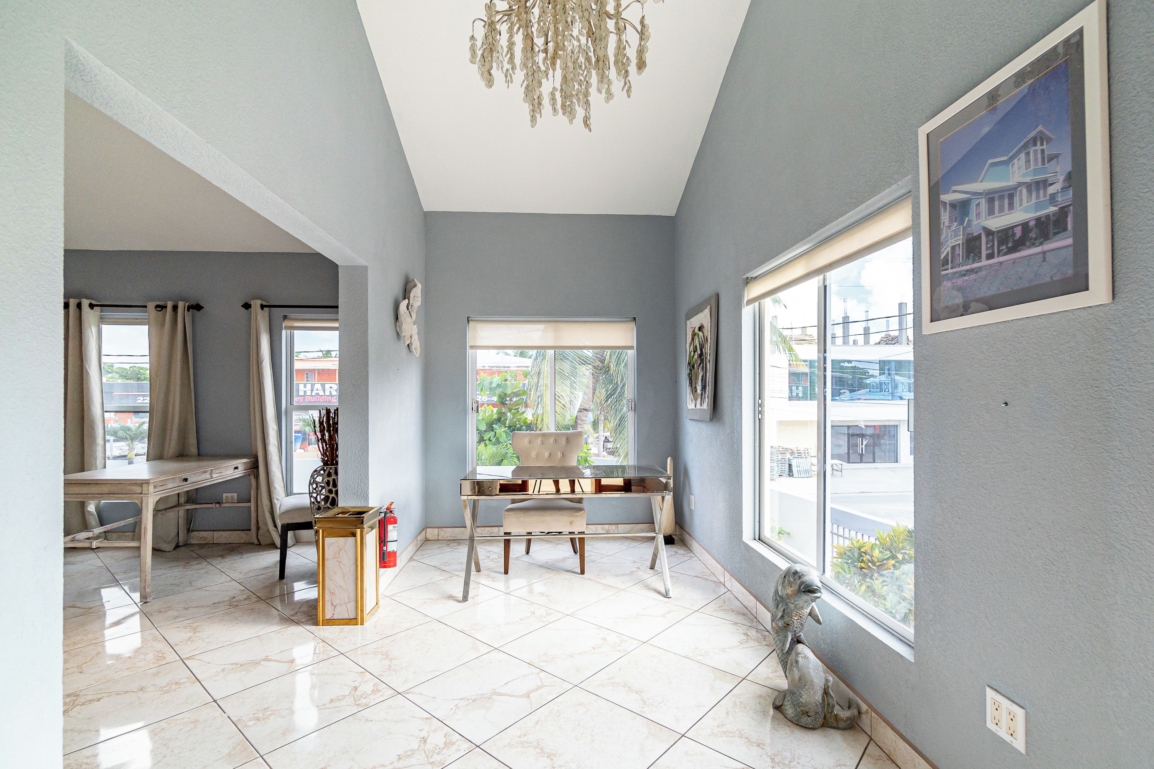 A spacious room with a dining table, chairs, a chandelier, and large windows offering a view of the outside.