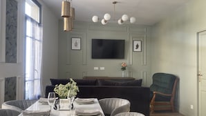 Un área de comedor con una mesa blanca dispuesta para cuatro personas, un sofá oscuro y un televisor montado en la pared.