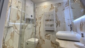Un baño con ducha de vidrio, inodoro blanco, pared con diseño de mármol, lavamanos blanco y un perchero para toallas.