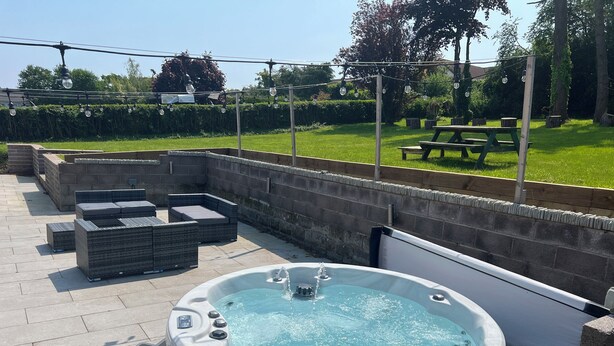 A hot tub surrounded by stone walls, outdoor seating, and a grassy area with trees and a picnic table. - Pine Lodge (Colwyn Bay)