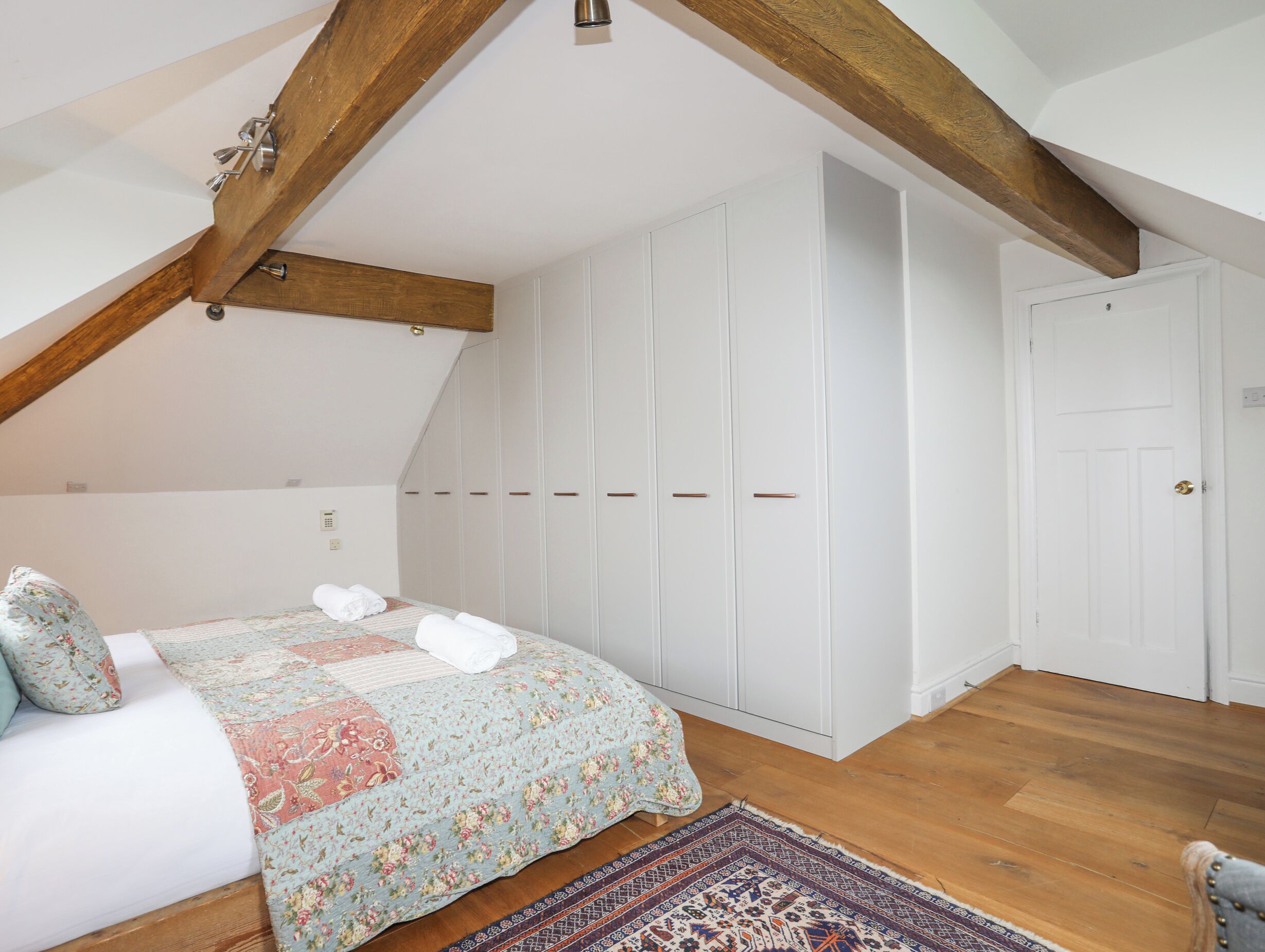 A bedroom with a wooden ceiling, a bed with floral bedding, a white wardrobe, and a patterned rug.
