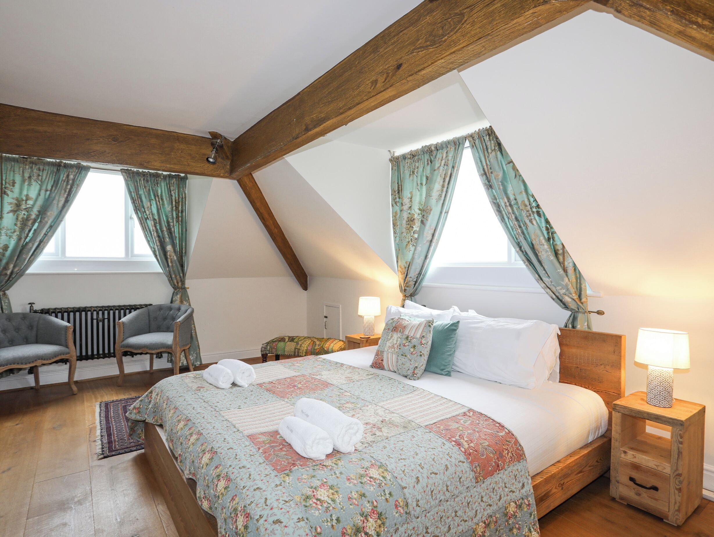 A cozy bedroom with a wooden ceiling, a large bed with floral bedding, two bedside tables with lamps, and a radiator.
