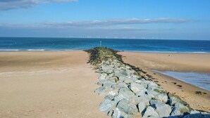 A beach with a rocky breakwater extending into the sea, a clear sky, and distant wind turbines. - Pine Lodge (Colwyn Bay)