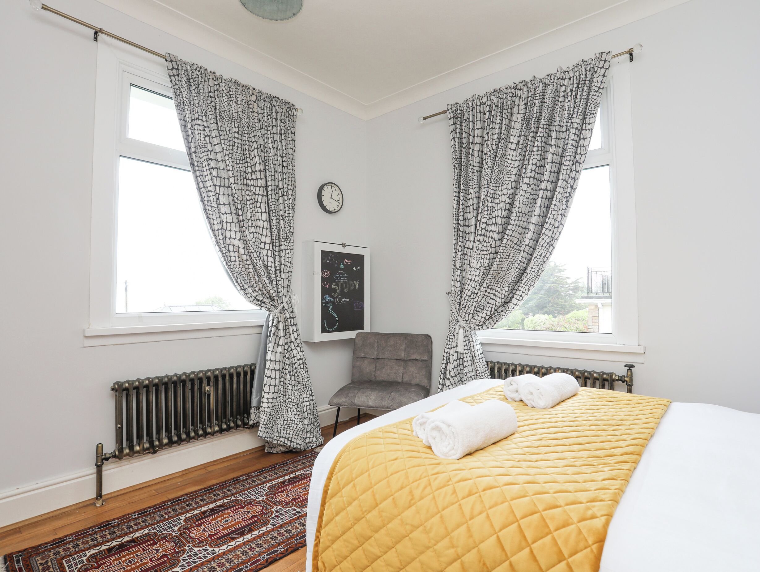 Une chambre à coucher avec un lit, deux fenêtres ornées de rideaux à motifs, un radiateur, une horloge et une petite chaise.