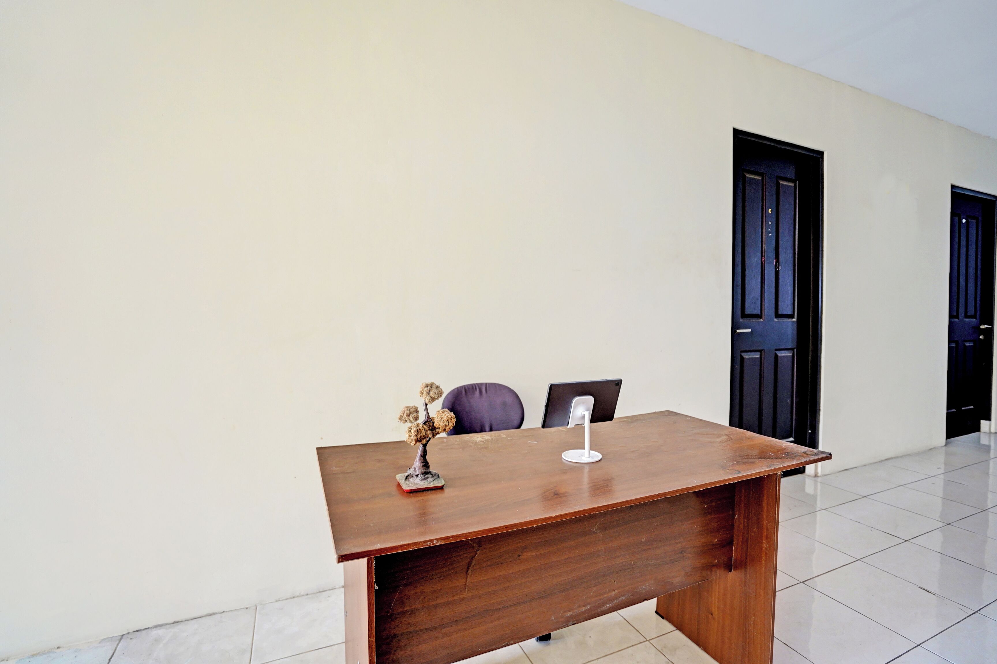 A wooden desk with a small decorative item, a chair, and a door in a room with a plain wall and tiled floor.