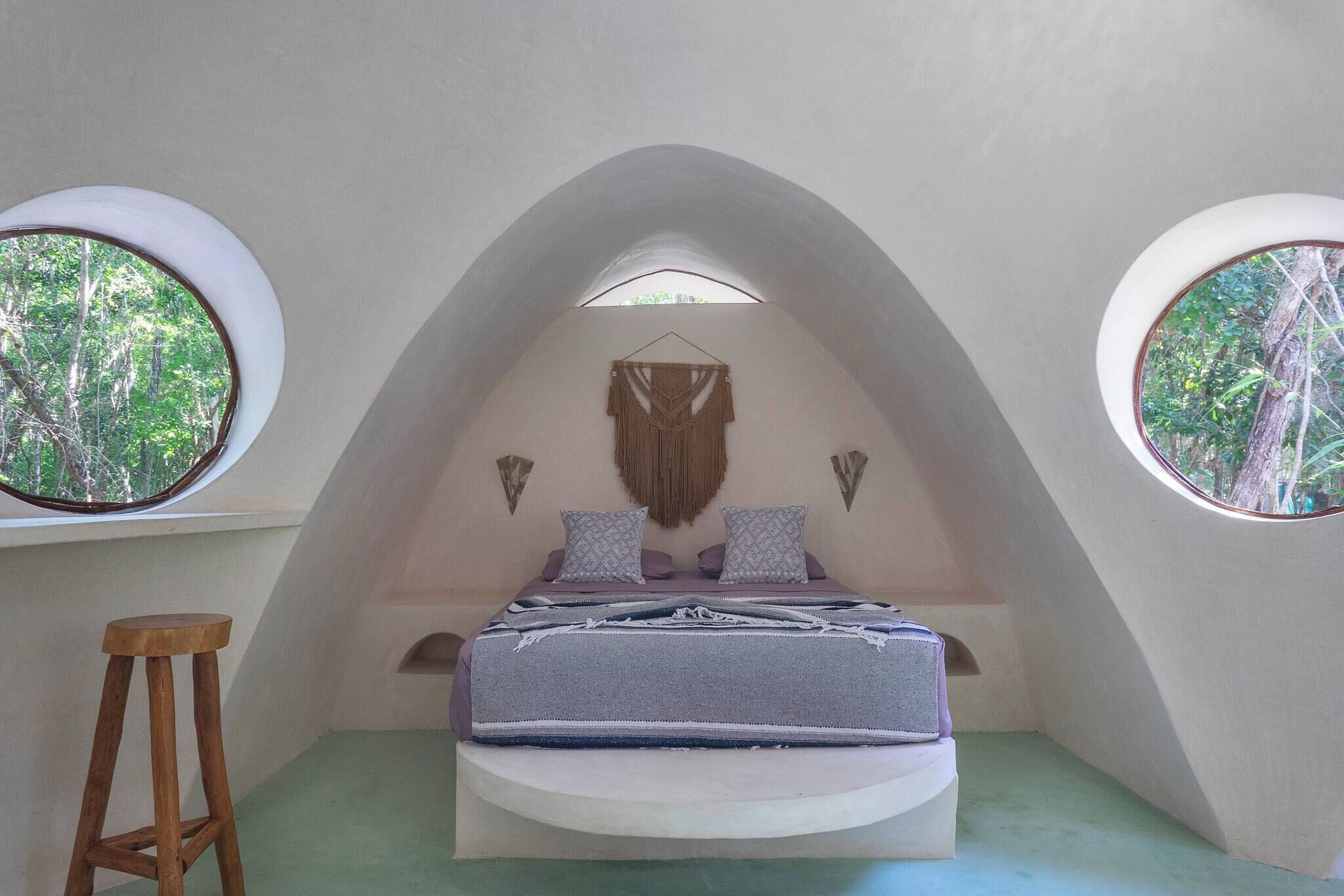 A bedroom with a bed, two windows, a hanging macrame piece, and a wooden stool.