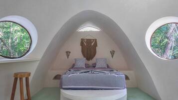 A bedroom with a bed, two windows, a hanging macrame piece, and a wooden stool.