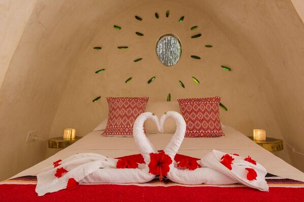 A cozy room with a bed, two pillows, a heart-shaped towel arrangement, and a circular mirror on the wall.