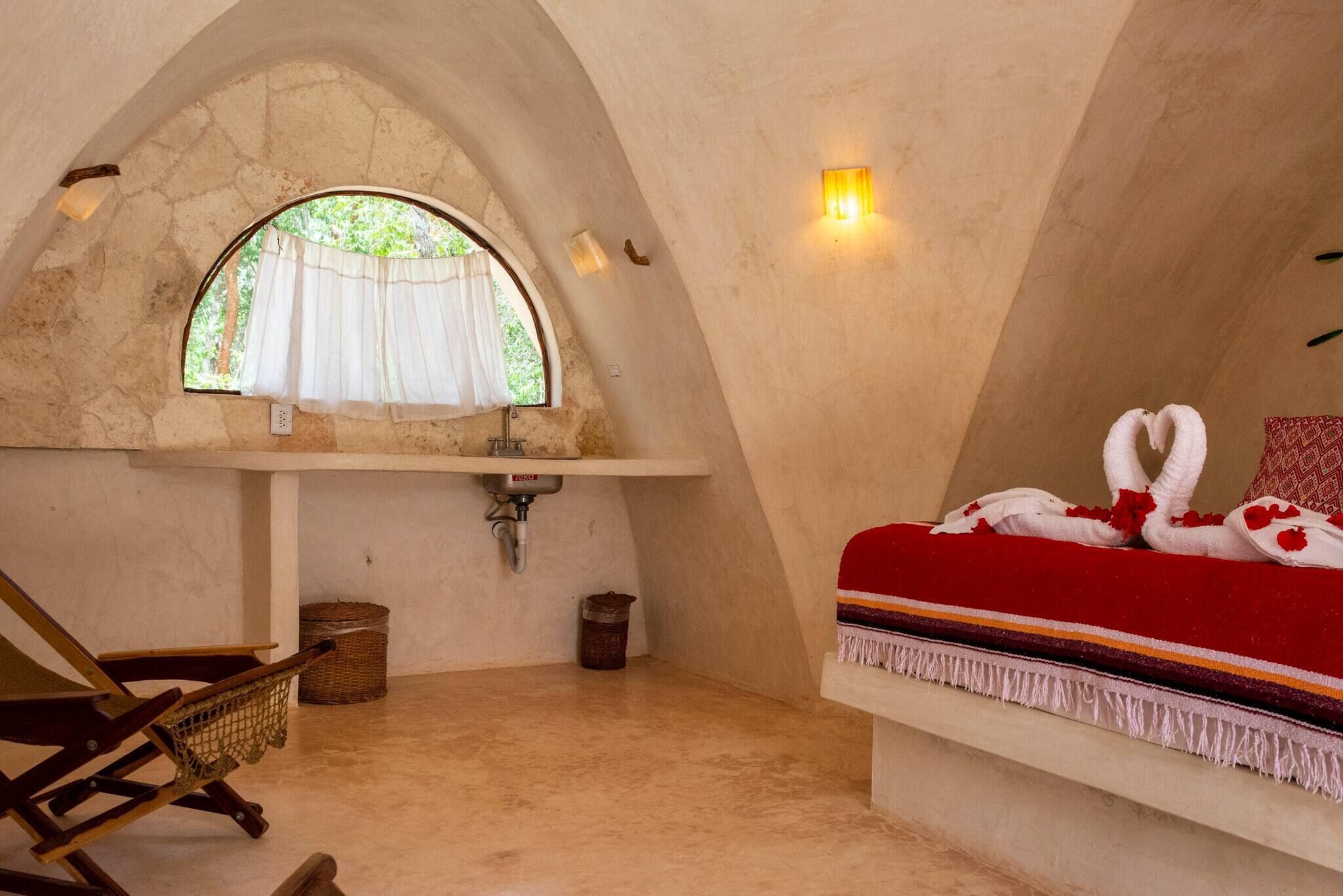 Una habitación acogedora con un ventanal de arco de piedra, una cama con manta roja, una silla de madera y un lavamanos pequeño.