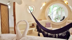 Un dormitorio acogedor con una hamaca, un ventanal redondo que ofrece vistas a la vegetación y una bañera blanca.