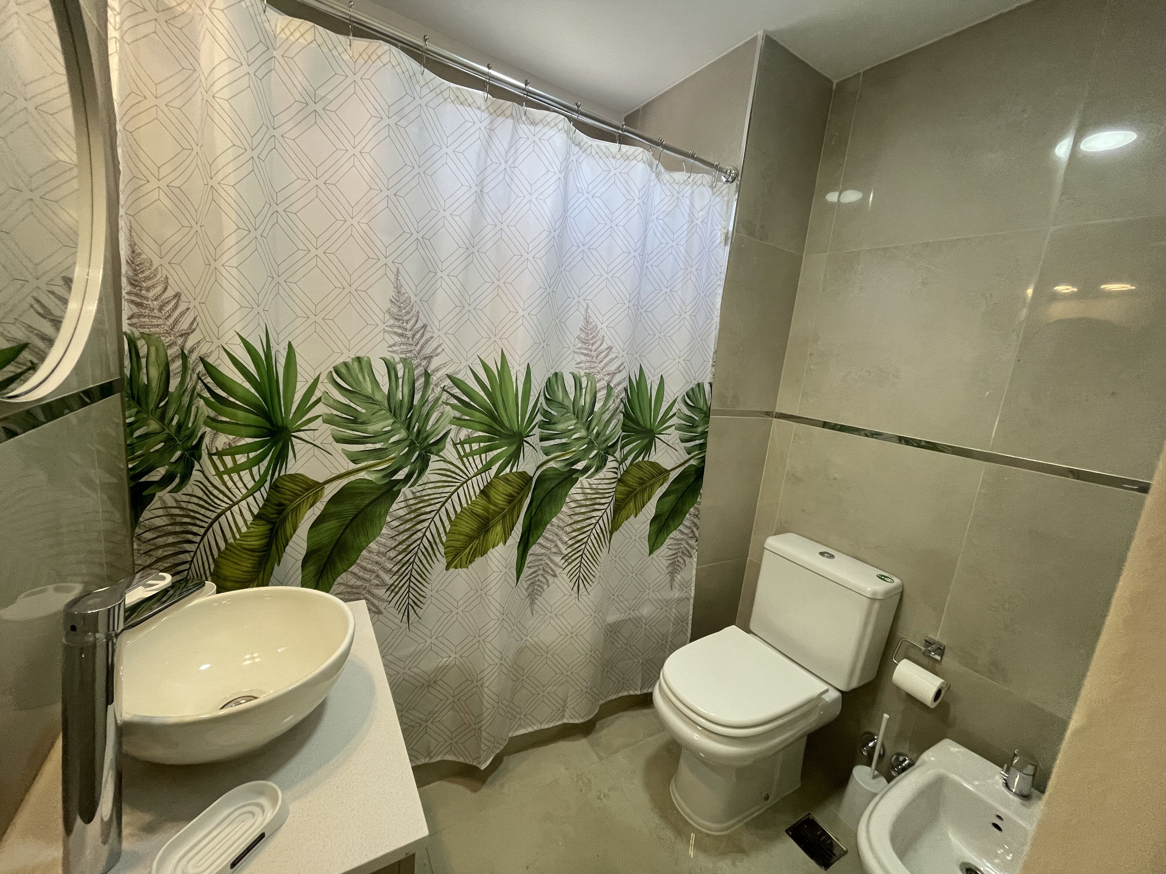 Un bany amb vàter, bidet, rentapel i cortina de dutxa amb un dibuix de fulles tropicals.