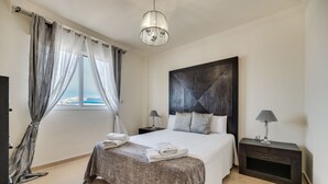 A bedroom with a large bed, a nightstand with a lamp, a window with curtains, and a view of the city.