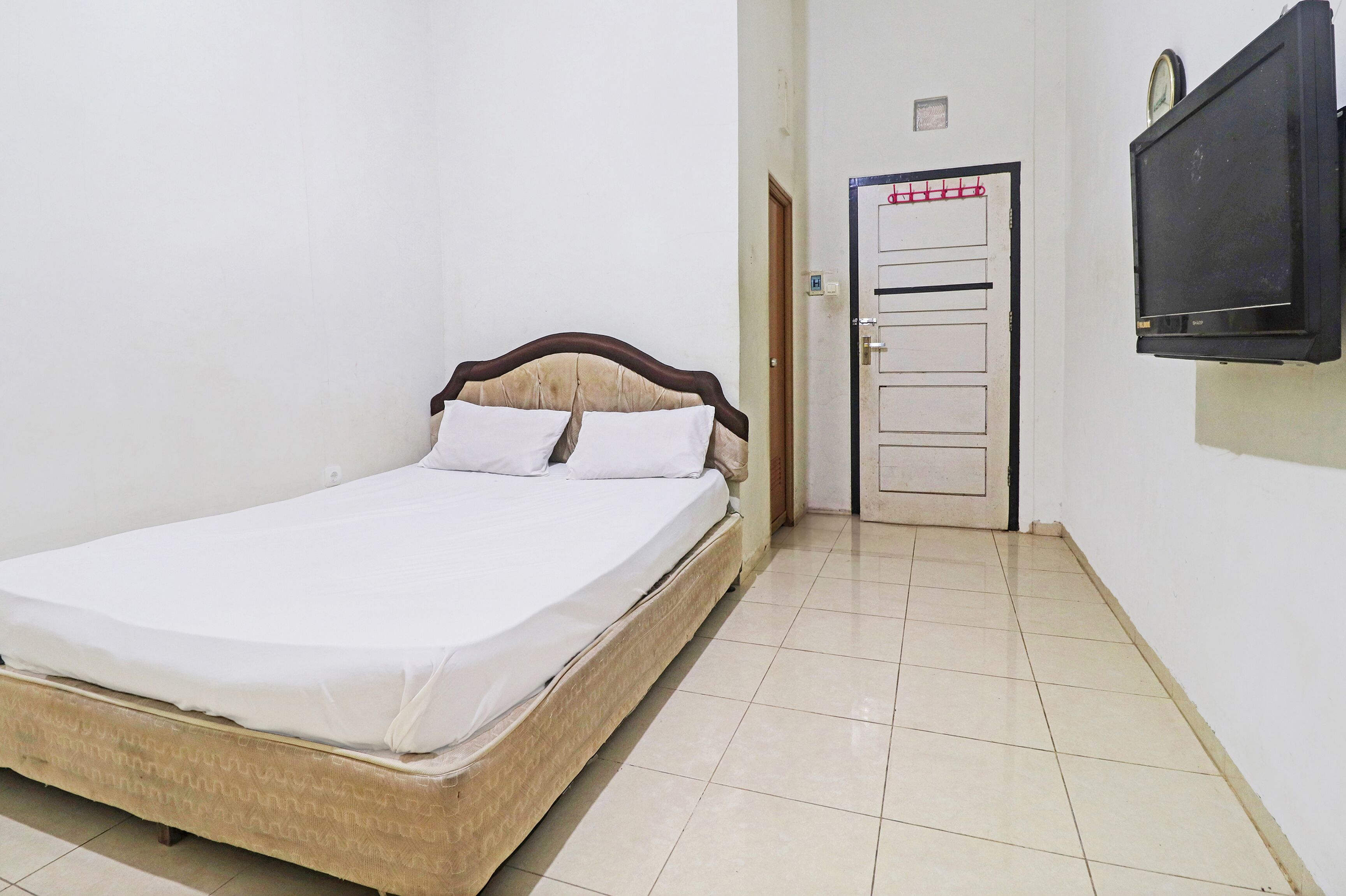 Une chambre avec un lit simple, une tête de lit en bois, un téléviseur fixé au mur et une porte avec une poignée rouge.