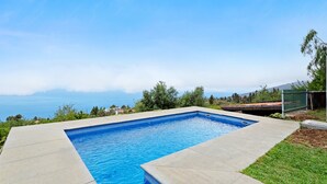 Ein rechteckiger Pool mit klarem, blauem Wasser, umgeben von einer Betonplattform, mit Grünflächen im Hintergrund und einem Blick auf das Meer in der Ferne.