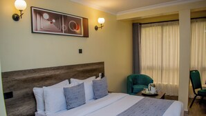 Een hotelkamer met een groot bed, een houten hoofdbord, twee wandlampen, een lijst met abstract schilderij, een groene fauteuil en een raam met dunne gordijnen.