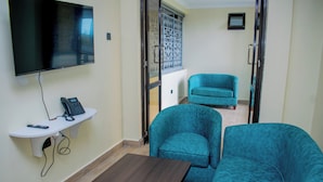 Een kamer met een wandgemonteerde flatscreen televisie, een klein wit schapje met een telefoon en afstandsbediening, een houten tafel en twee teal fauteuils.