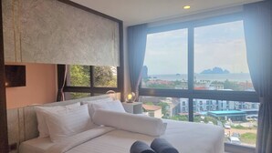 A hotel room with a large bed, a nightstand, a lamp, and a view of the city and sea.