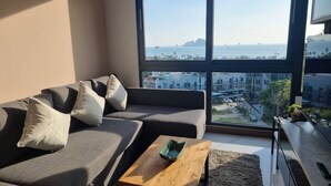 Ein modernes Wohnzimmer mit einem grauen Sofa, einem Holztisch und einem großen Fenster mit Blick auf das Meer und die Stadt.
