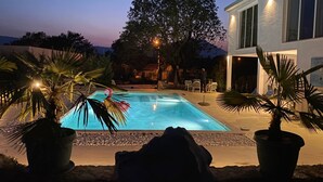 A swimming pool with a stone border, surrounded by potted plants, and a building with large windows in the background. - The Rock Star's Villa With Private Pool And Beach (Novo Selo)