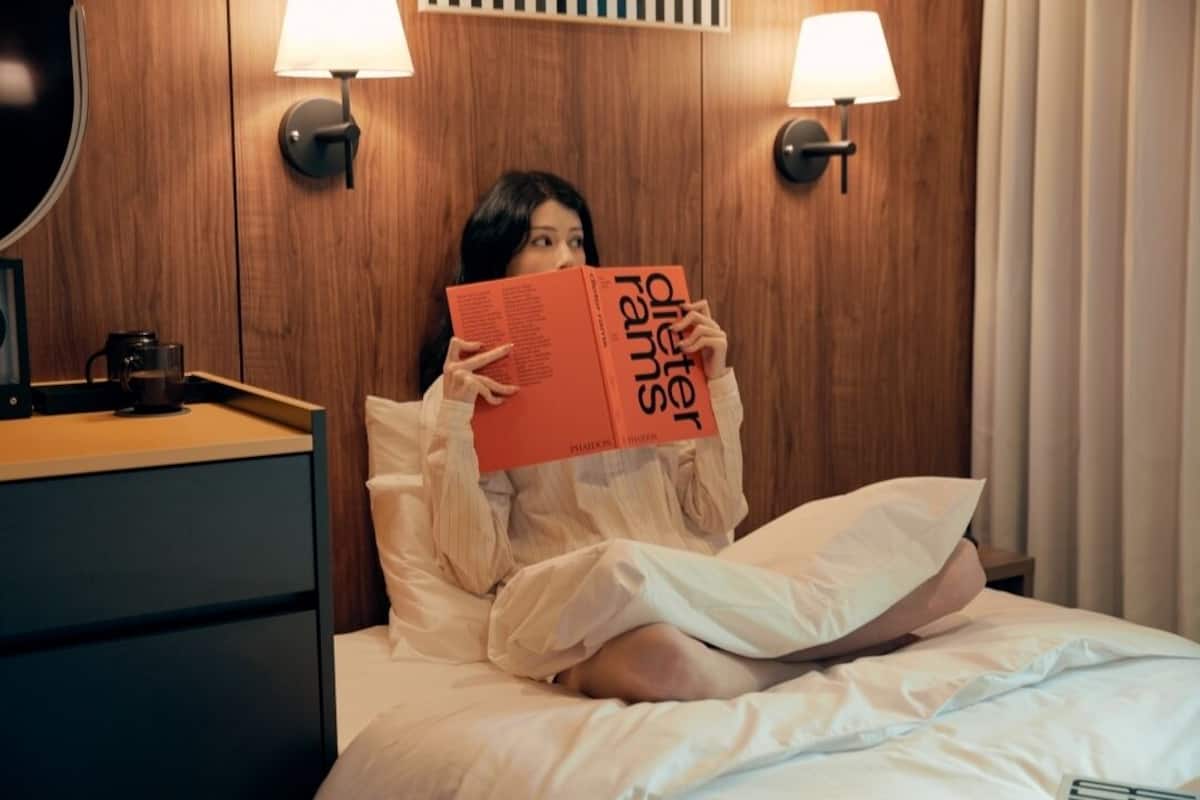 Una persona leyendo un libro en una habitación de hotel con paneles de madera, dos lámparas de noche y una mesita de noche con una cafetera.