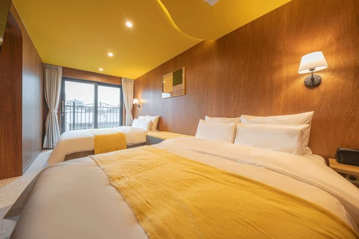 Habitación de hotel con dos camas, paredes de madera, un ventanal grande con cortinas y una lámpara fijada a la pared.