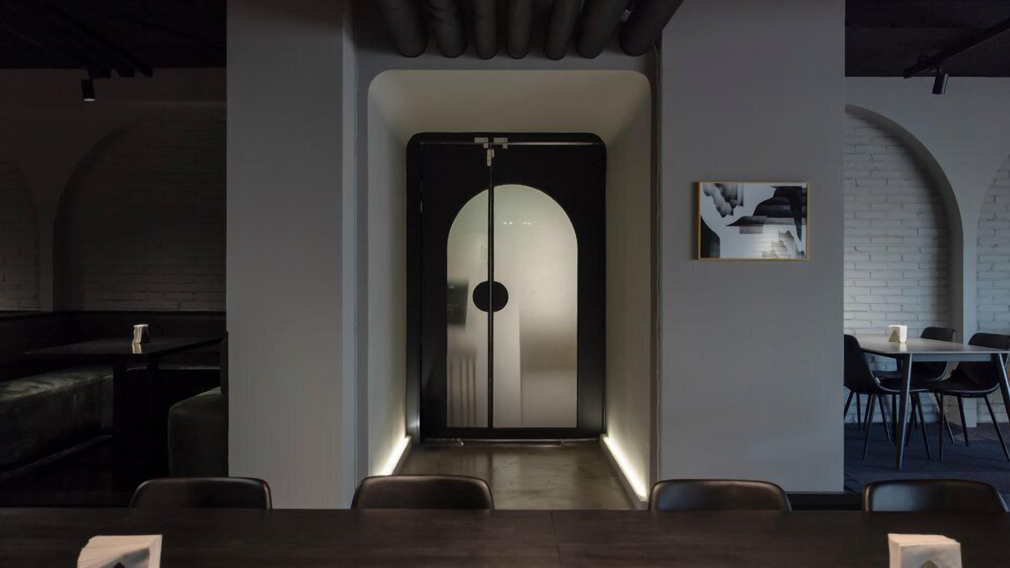 A modern interior with a central door leading to a dark room, a seating area with tables and chairs, and a brick wall with a framed picture.