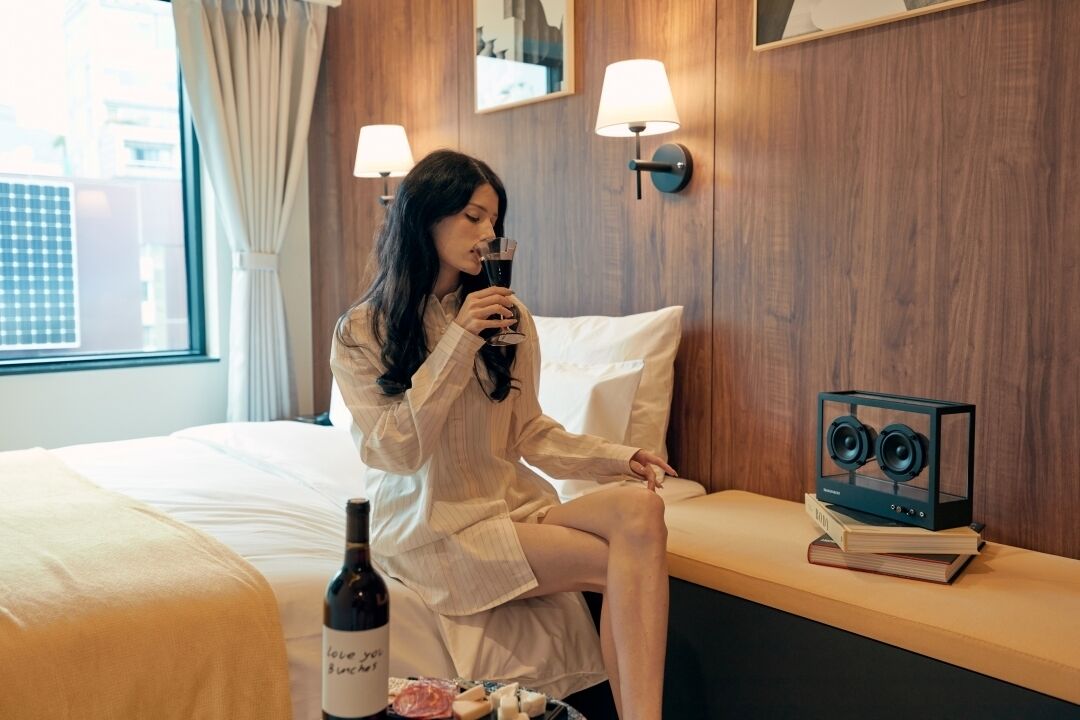 A person sitting on a bed in a hotel room with a bottle of wine, a book, and a speaker on a nightstand.