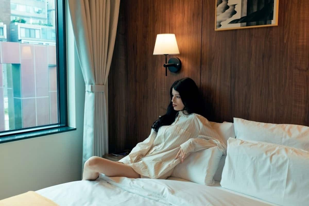 Una persona sentada en una cama en una habitación con pared de madera y una ventana con cortinas.