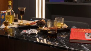 Una habitación con una pared oscura, un espejo con luz de neón, un libro titulado "Art =", y varios objetos sobre una encimera, incluyendo una botella, vasos y un libro.