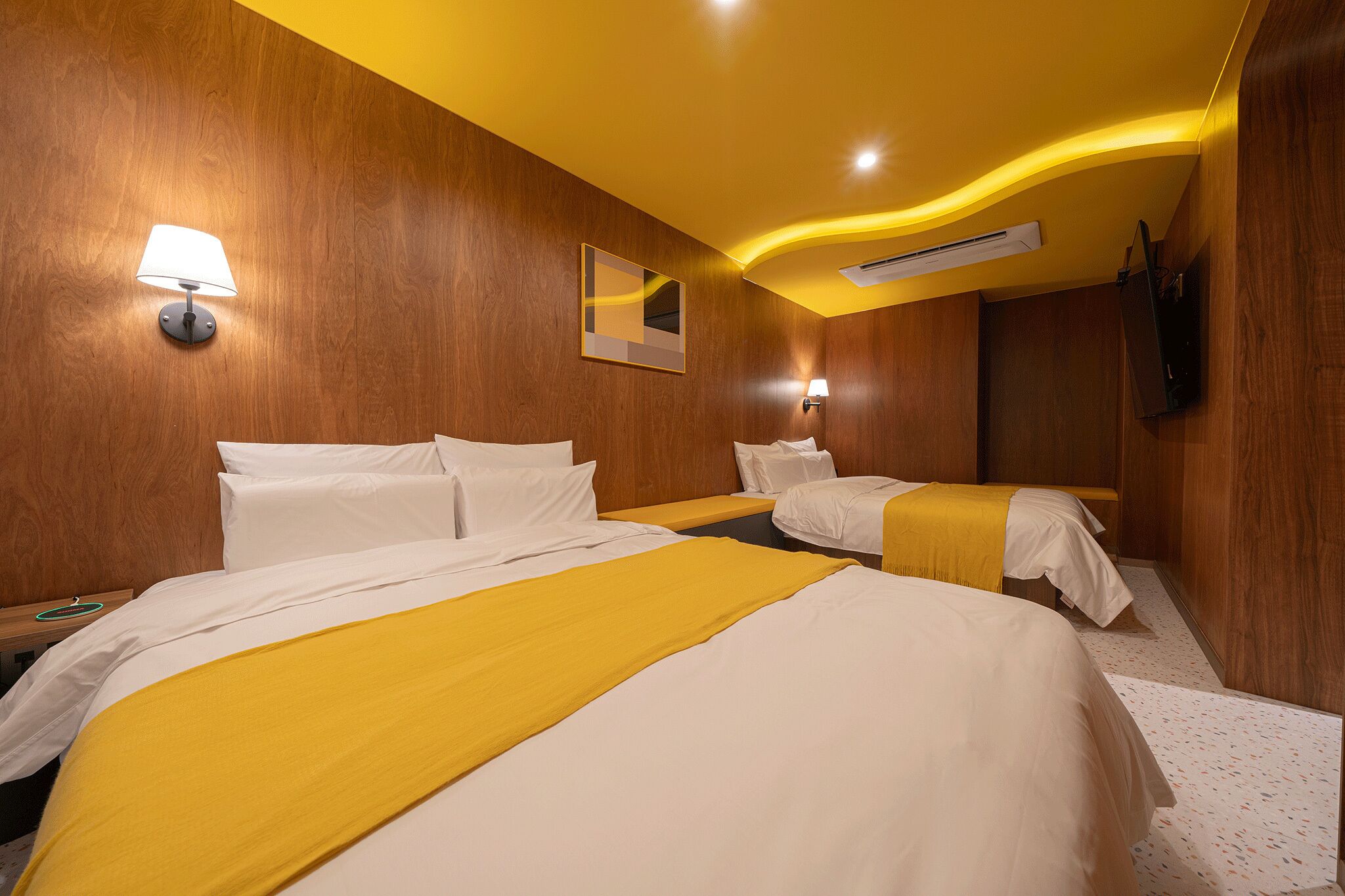 Habitación de hotel con dos camas, paredes de madera, un espejo, televisor fijado a la pared y una lámpara en el techo.