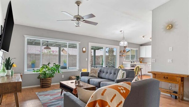 Ein geräumiges Wohnzimmer mit grauem Sofa, einem Couchtisch und einem Fernseher auf einem Holzbodenständer. Ein Deckenventilator, große Fenster und ein Blick in den Küchenbereich sind ebenfalls zu sehen.