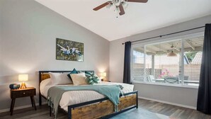 Ein Schlafzimmer mit Bett, Nachttischen, Deckenventilator, einem Fenster mit Vorhängen und Blick ins Freie.
