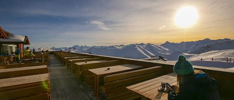 Une terrasse en bois avec des bancs, d’où l’on peut admirer un paysage montagneux enneigé au coucher du soleil.