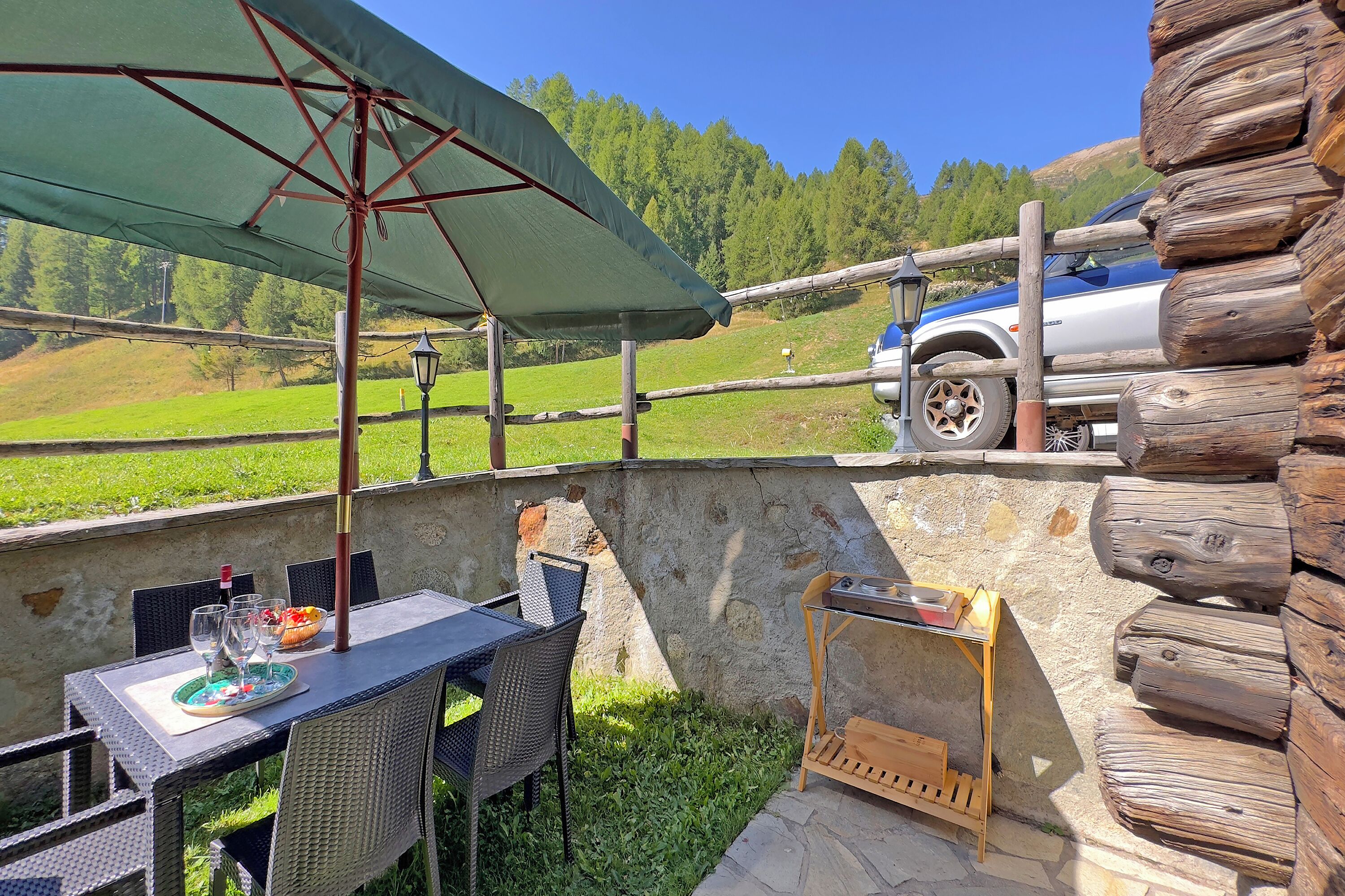 A patio with a table, chairs, and a grill, overlooking a green landscape and a parked car.
