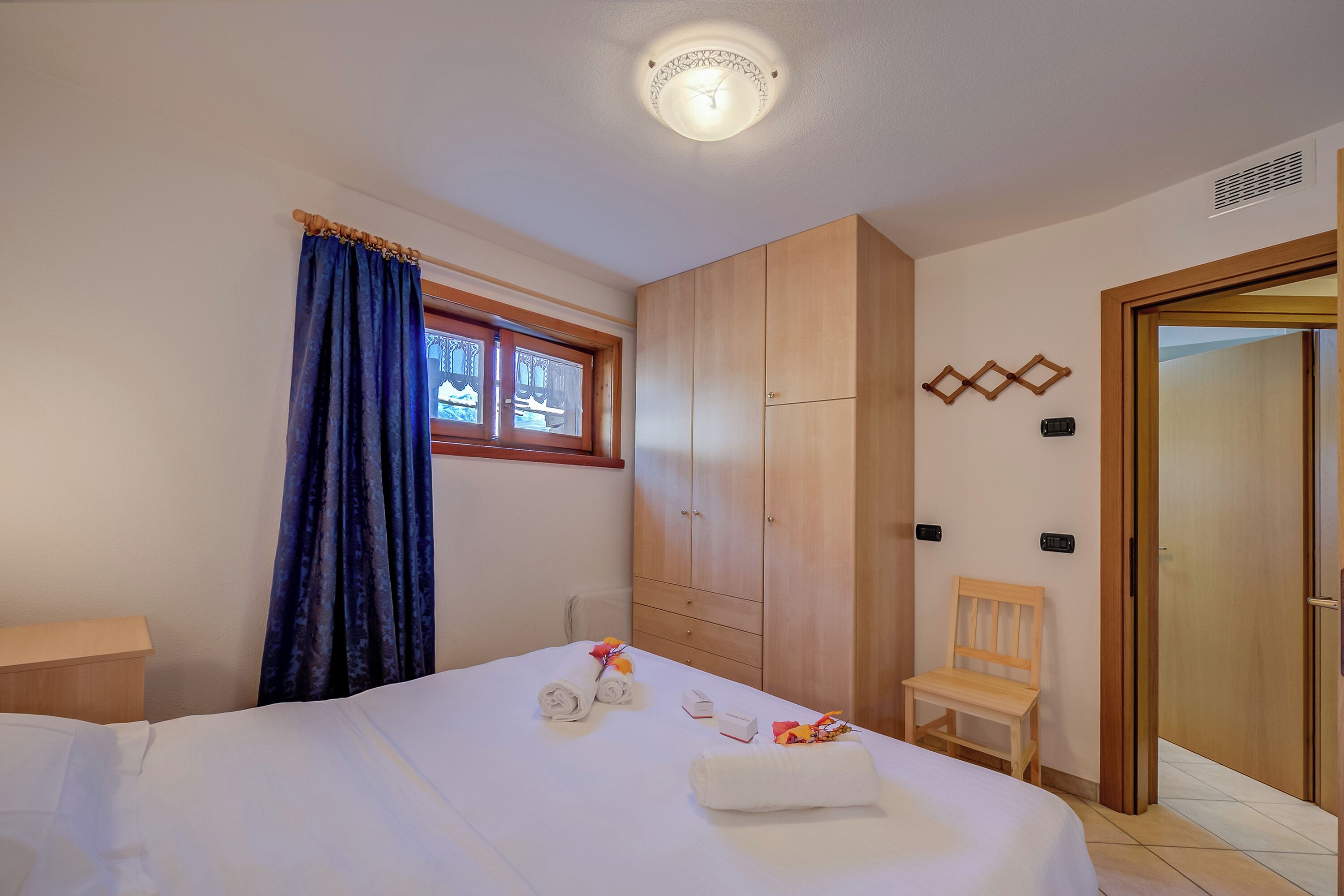 A hotel room with a bed, a wooden wardrobe, a window with curtains, a wooden chair, and a door.