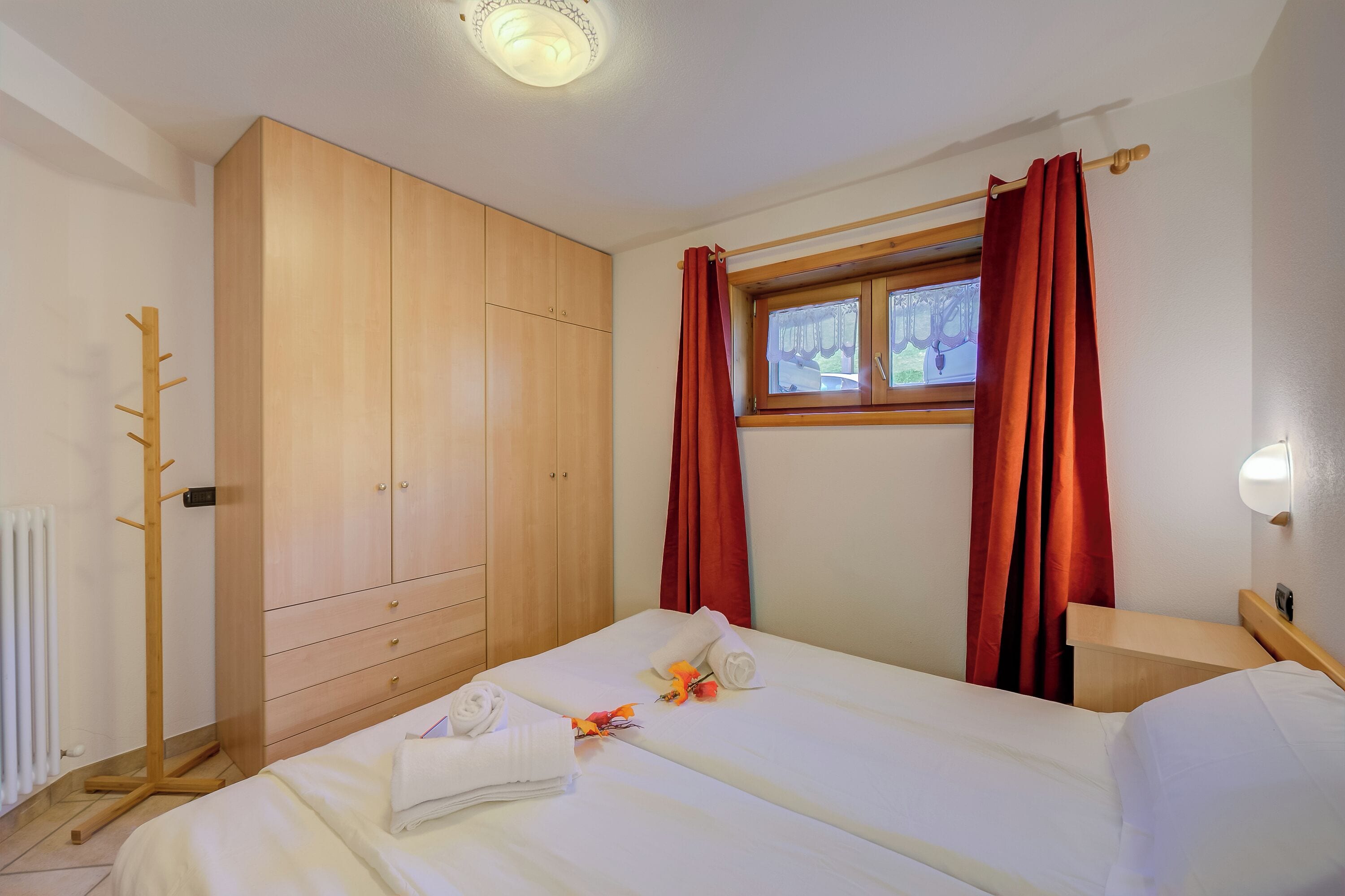 A hotel room with a bed, a wooden wardrobe, a window with red curtains, and a wooden nightstand.