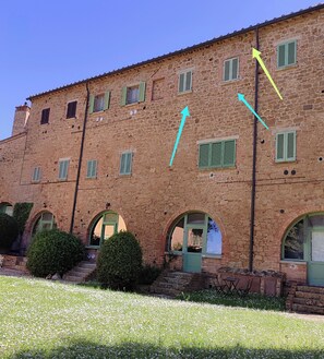 A stone building with multiple windows and green shutters, featuring a courtyard with a grassy area and a small garden.