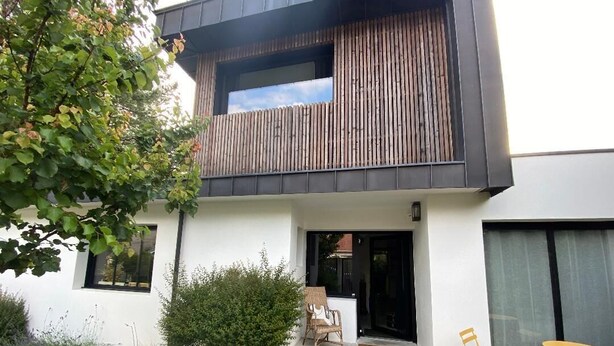 A modern two-story building with a balcony, a small outdoor seating area, and a door leading to the entrance.