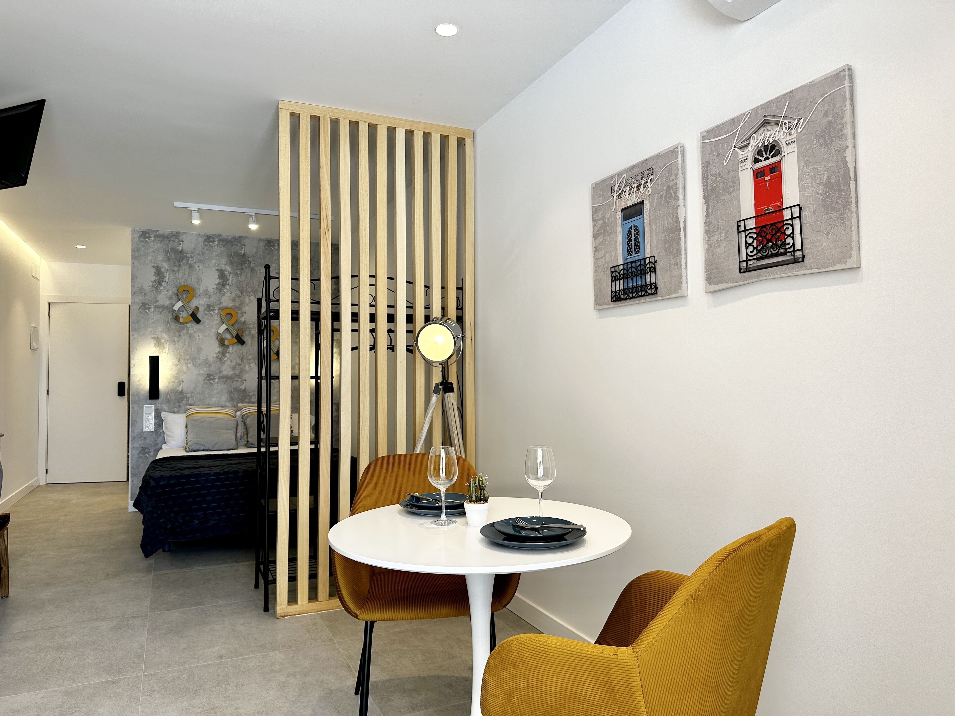 A modern hotel room with a bed, a dining area with a table and chairs, a decorative wall with artwork, and a sliding door.