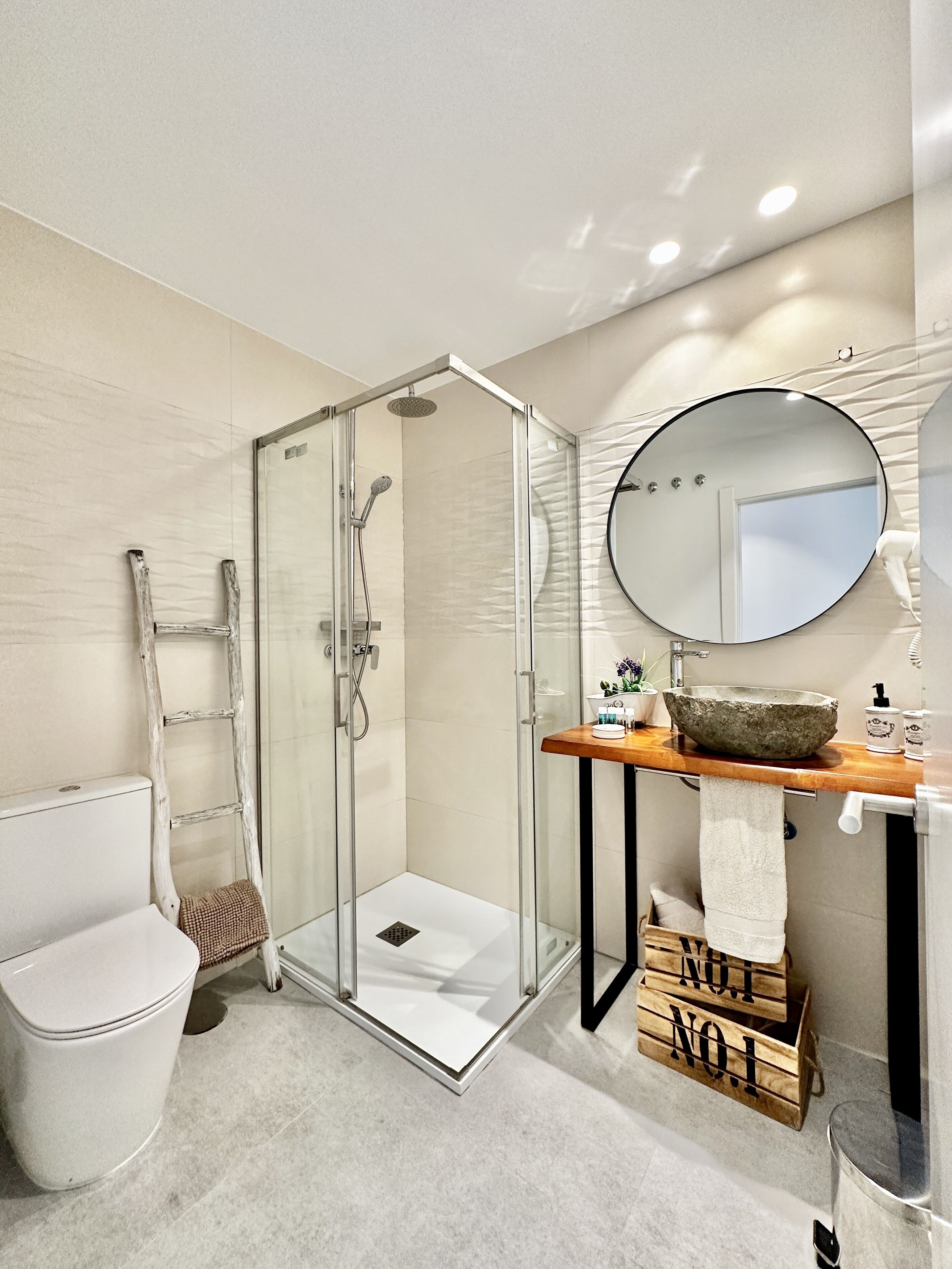 A modern bathroom with a glass shower enclosure, a wooden vanity with a stone basin, and a round mirror.
