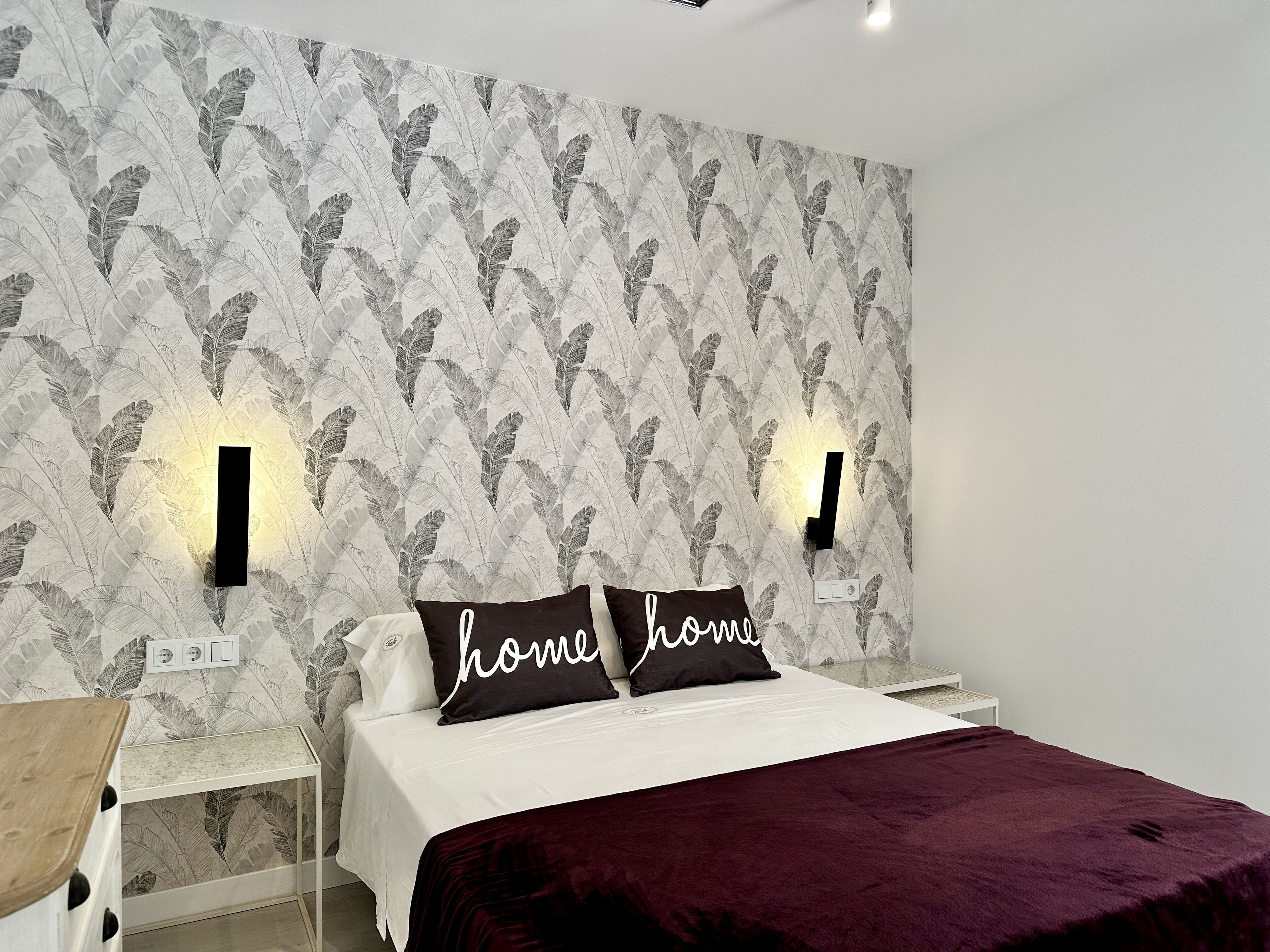 A bedroom with a patterned wallpaper, two bedside lamps, a bed with a dark red blanket, and a nightstand with a lamp.