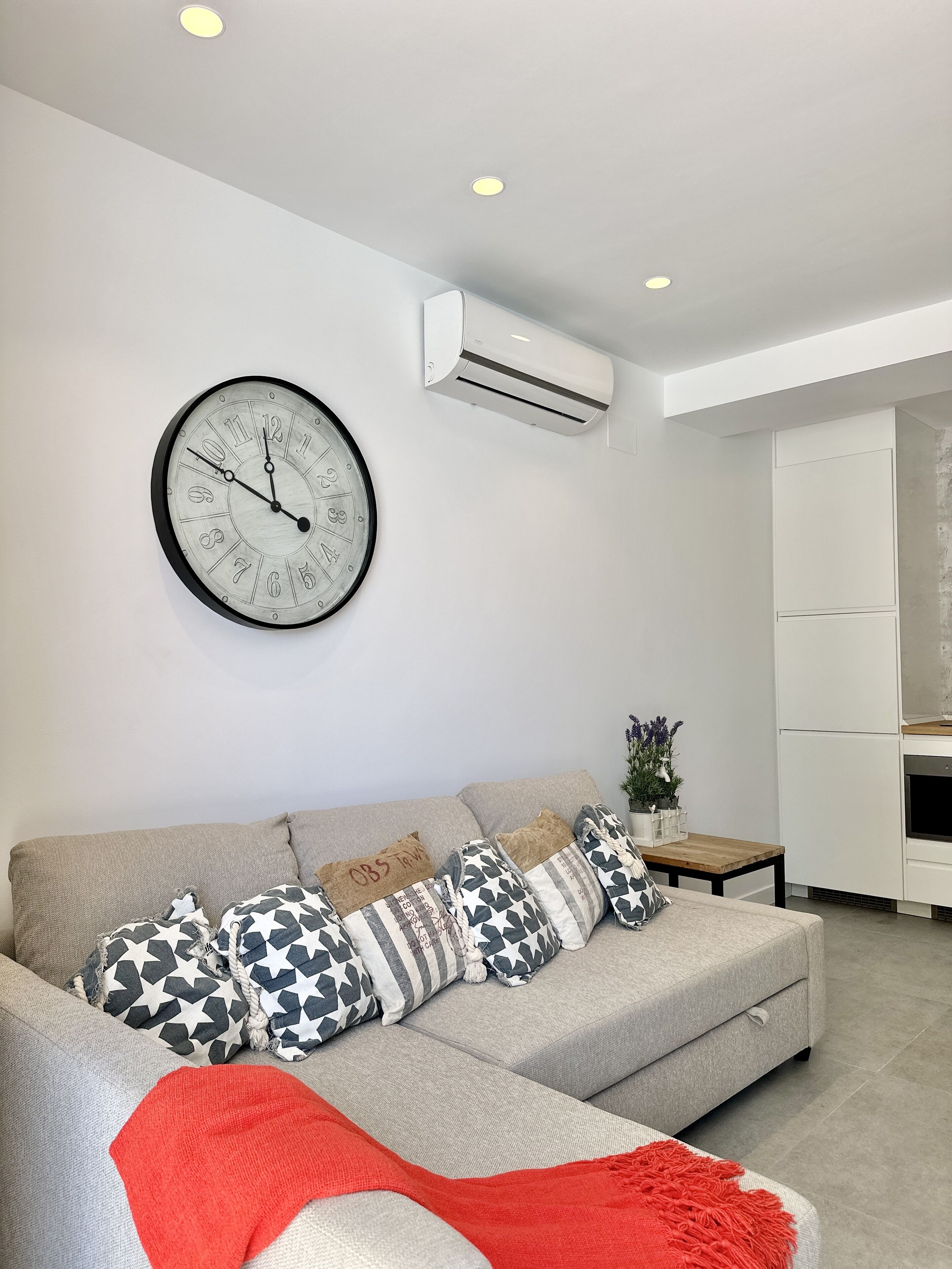 A modern living room with a grey sofa, patterned cushions, a red throw blanket, a wall clock, and an air conditioning unit.