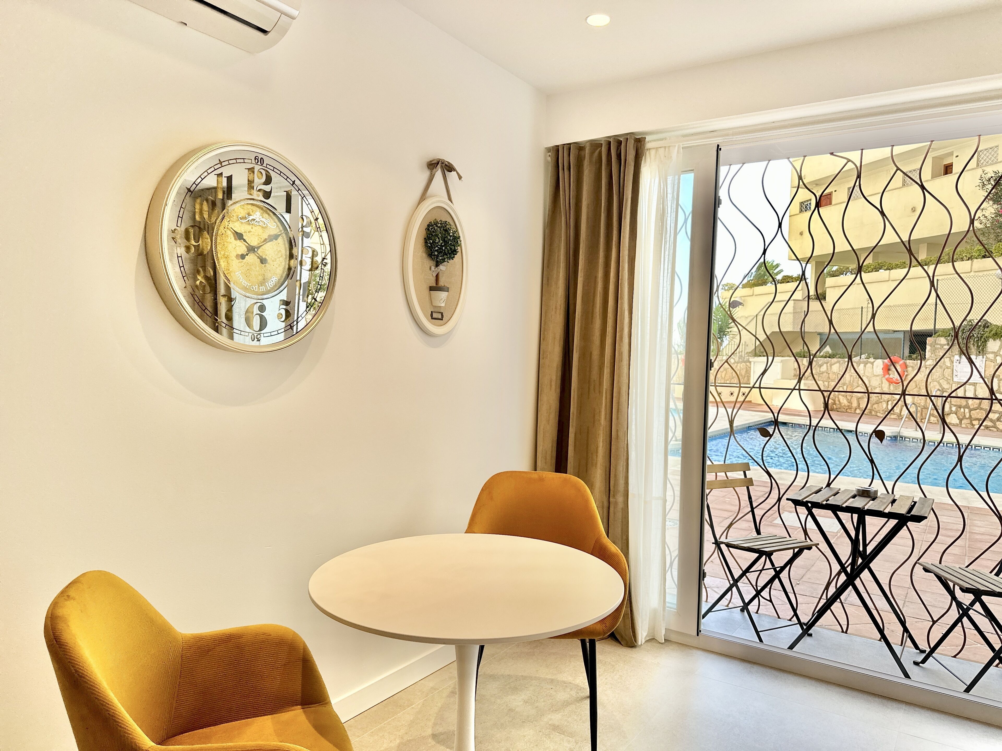 Una habitación con un ventanal grande, una mesa redonda, dos sillas color mostaza y dos relojes de pared.